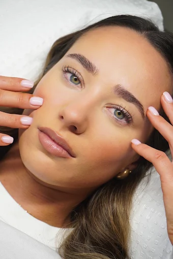 A client resting with hands framing the face, showing brows, lashes and pale pink nails together.
