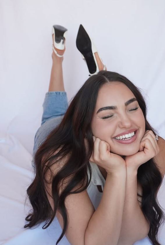 A woman lying on white linen, laughing, heels kicked up behind her.