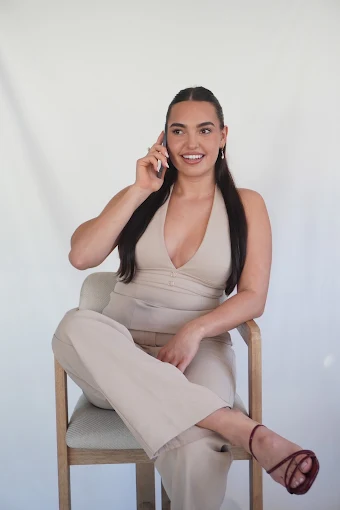 A woman seated on a timber chair against a plain backdrop, on the phone and smiling.