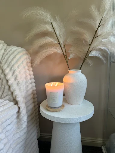 A lit candle and dried pampas in a white vase on a small round side table.