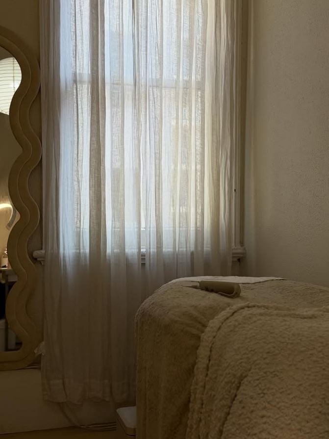 The treatment room, with sheer linen curtains at a tall window and a bed dressed in cream.